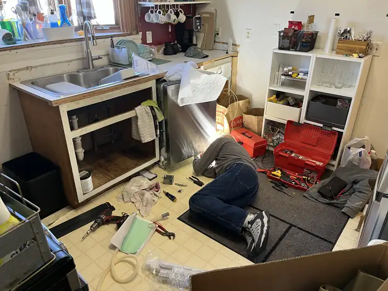Technician lying on a kitchen floor working inside the cabinet under the sink, with a red toolbox beside him